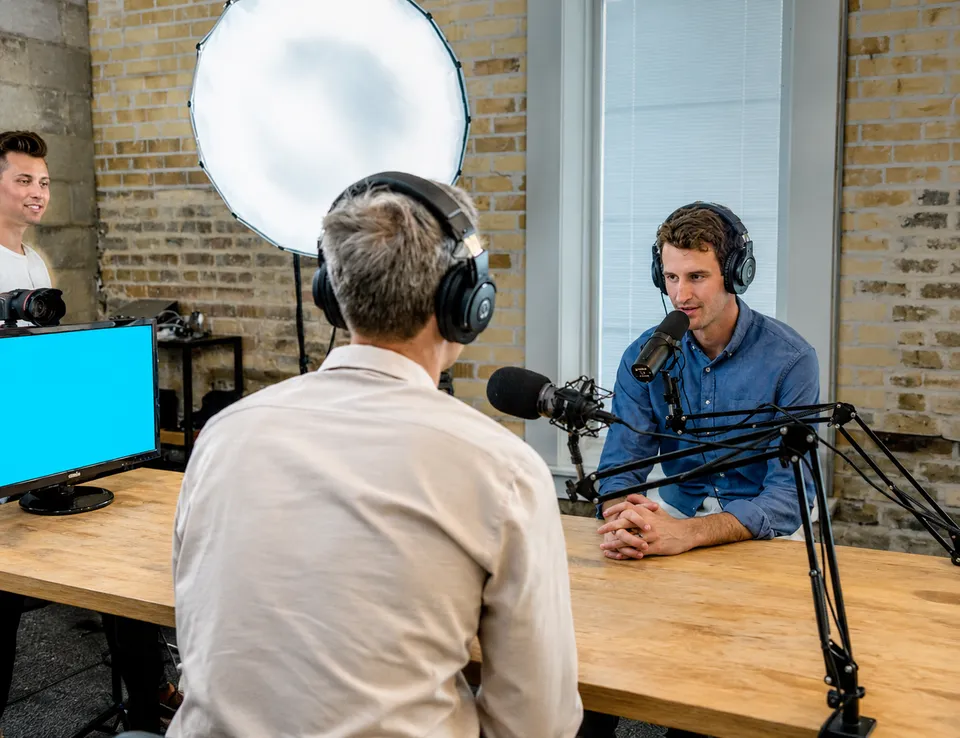 Podcast host interviewing a guest in a studio with boom-arm mics and a softbox light